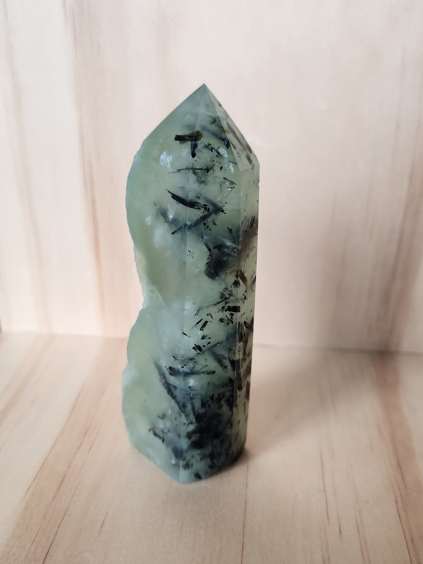 May include: A green crystal point with black inclusions. The crystal is smooth and polished with a pointed tip. The crystal is a natural formation with unique markings and patterns.