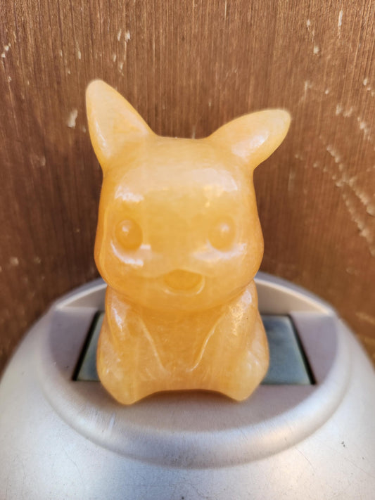 May include: A small, light orange, Pikachu figurine carved from polished gemstone. The figurine is sitting on a light gray circular base.  The Pikachu is detailed with simple features, including round eyes and a small, open mouth. The smooth, translucent stone gives the figurine a warm, glowing appearance.
