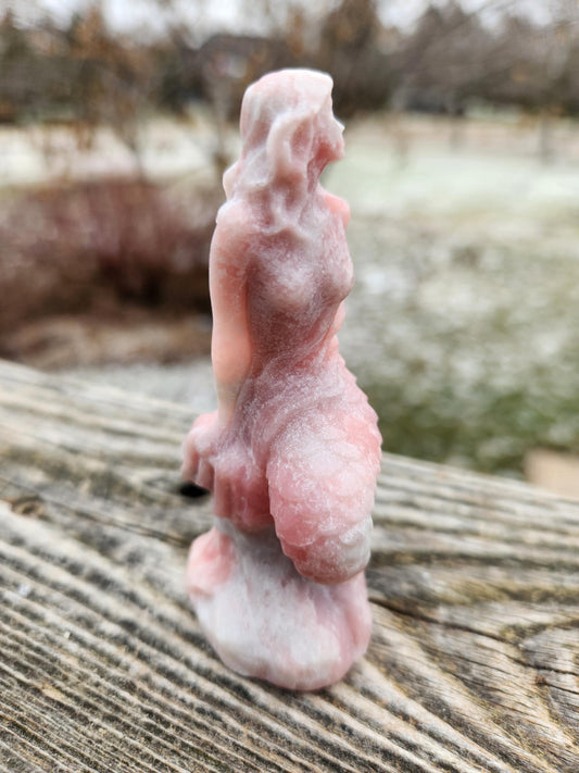 May include: A pink and white mermaid figurine made of stone. The figurine is standing on a wooden surface.