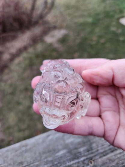 May include: Clear quartz carved into the shape of a detailed three-dimensional toad. The toad's body is covered in small, bubble-like textures.  Intricate carvings adorn the toad's face and body, showcasing fine craftsmanship. This collectible figurine is a feng shui symbol of wealth and prosperity, perfect for home decor or as a gift.