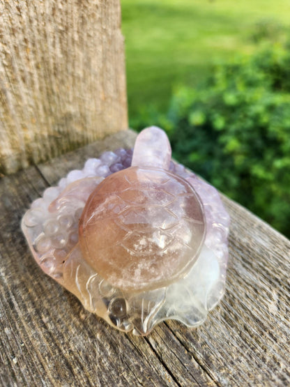 May include: A carved crystal turtle figurine with a pink and purple color gradient. The turtle is sitting on a wooden surface.