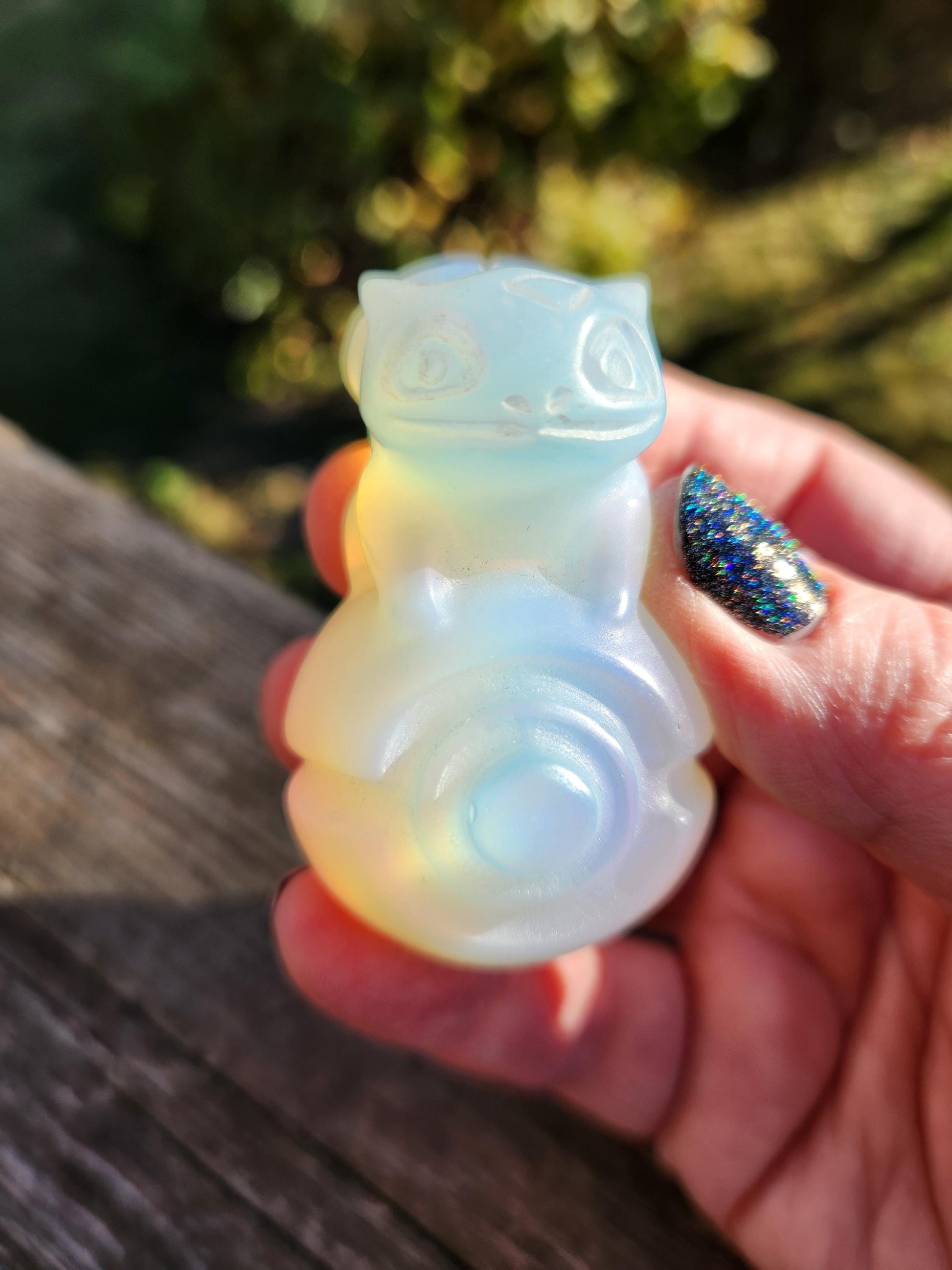 May include: A pale, translucent opalite carving of a mythical creature, possibly a Pokémon, sits on a dark brown wooden surface. The creature is light blue and white, with a rounded body and a small, stylized head. The carving is smooth and polished, with subtle color variations throughout.  The opalite shows a soft, ethereal glow. This collectible figurine is a unique piece of fantasy-themed decor.