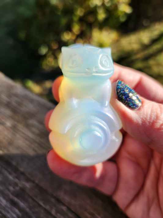 May include: A pale, translucent opalite carving of a mythical creature, possibly a Pokémon, sits on a dark brown wooden surface. The creature is light blue and white, with a rounded body and a small, stylized head. The carving is smooth and polished, with subtle color variations throughout.  The opalite shows a soft, ethereal glow. This collectible figurine is a unique piece of fantasy-themed decor.