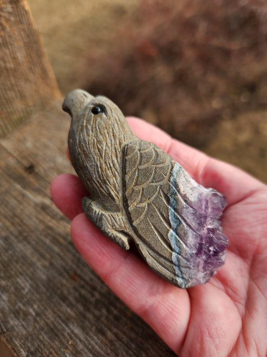 May include: A carved eagle figurine made of amethyst with a purple and gray color scheme. The eagle is perched with its wings spread and has a detailed carving of its feathers.