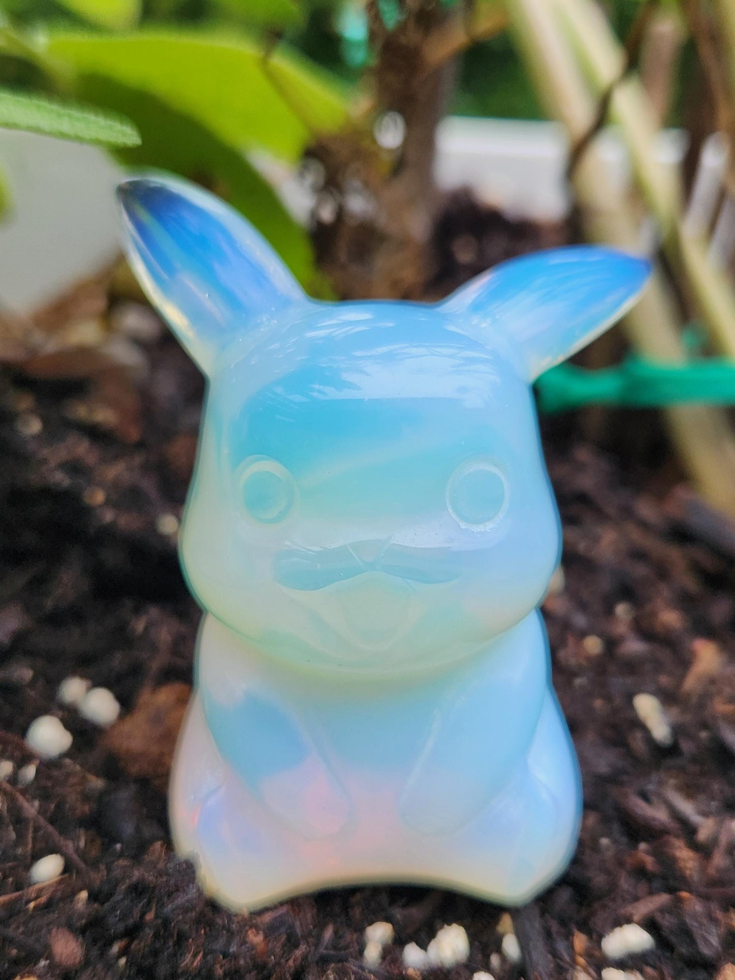 May include: A Pikachu figurine made of opalescent glass. The figure is in a sitting pose with its arms at its sides. The ears are pointed and blue-tipped. The body is a light blue with a white belly and a small, smiling mouth. The figure is set against a background of dark soil and green foliage.